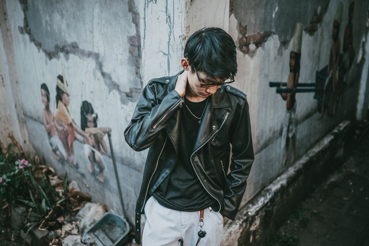 Stylish young man in a leather jacket stands against a mural wall.