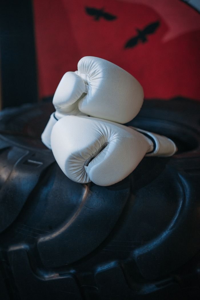 Close-up of white boxing gloves resting on a large tire in a gym setting.